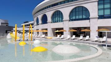 A modern hotel with a pool area featuring sun loungers and umbrellas, a large building with arched windows, and a clear blue sky.