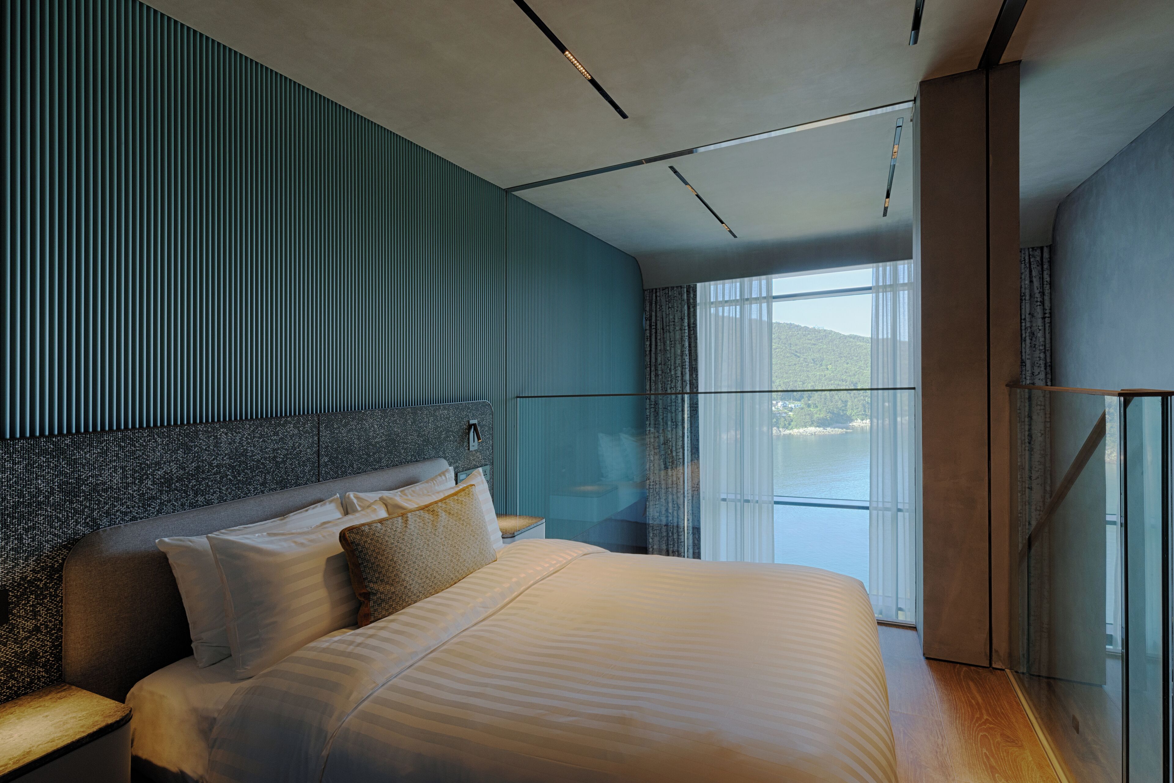 Habitación de hotel moderna con una cama grande, cabecera con líneas verticales, mesita de noche y vista a un cuerpo de agua a través de una puerta de cristal.