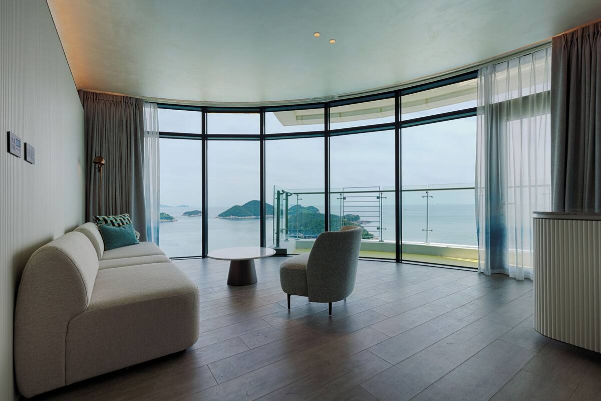 A modern living room with a curved glass wall, a sofa, a chair, and a small table.