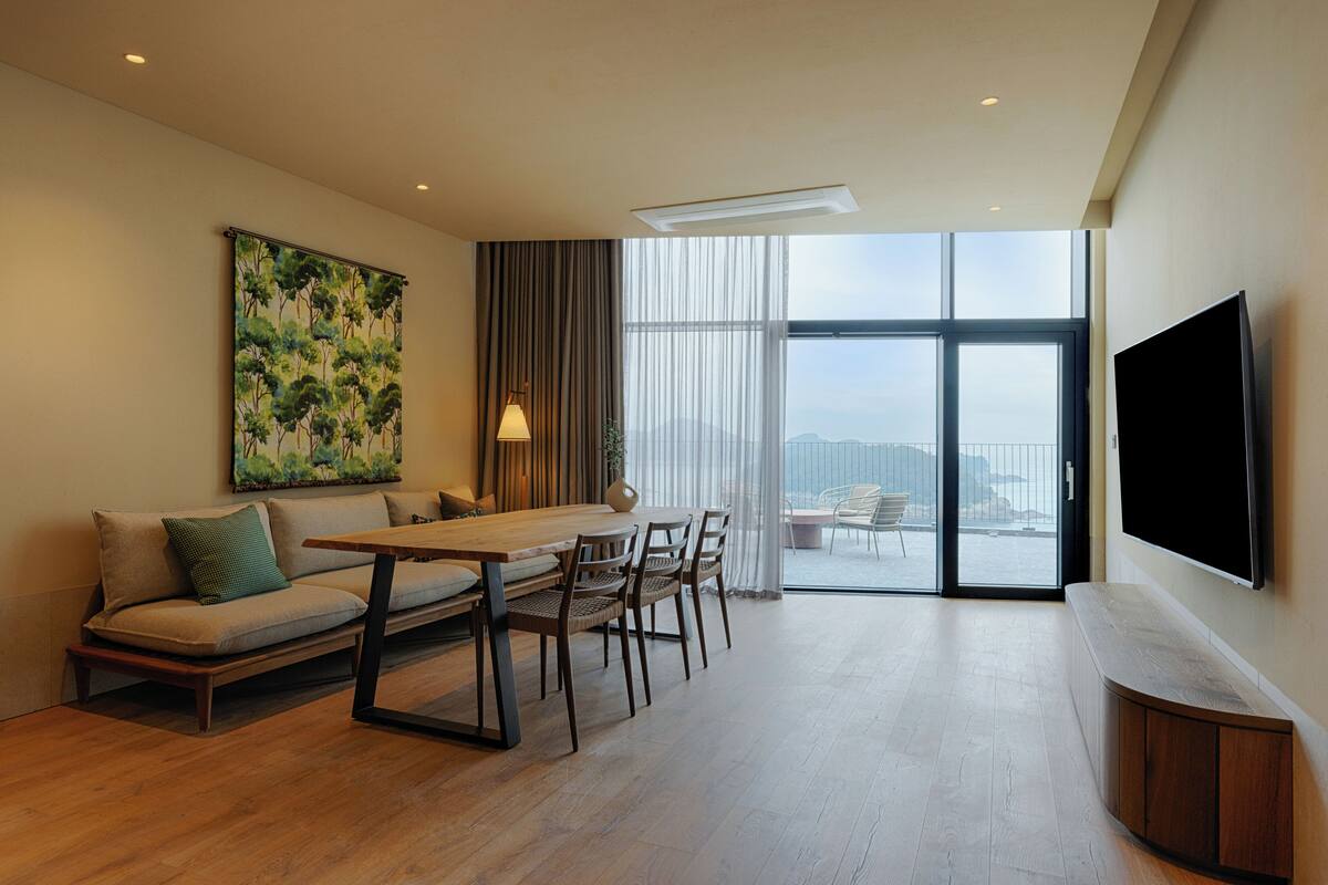 A modern living room with a sofa, dining table, chairs, a TV, and a large window offering a view.