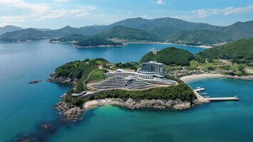 Vista aérea de un resort costero con una estructura hotelera moderna, rodeado de exuberante vegetación y montañas.