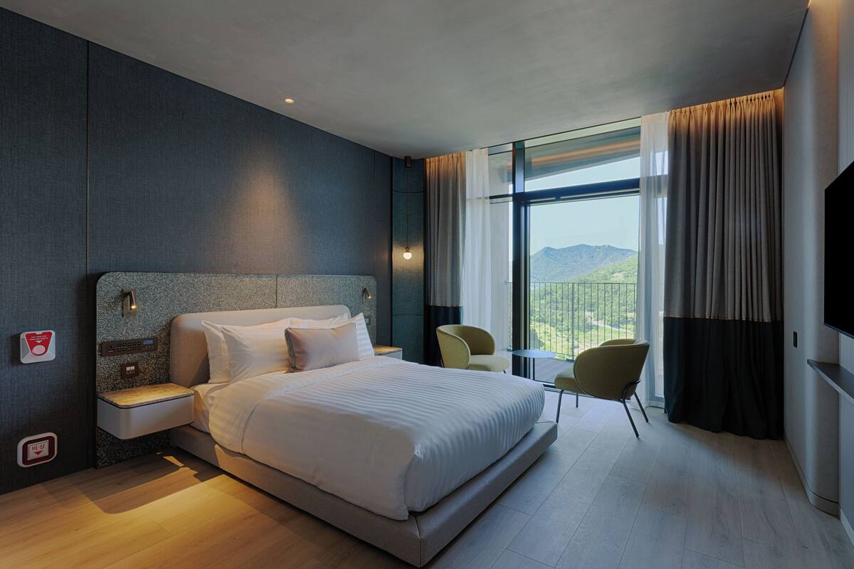 A modern hotel room with a large bed, bedside tables, a TV, and a view of the mountains.