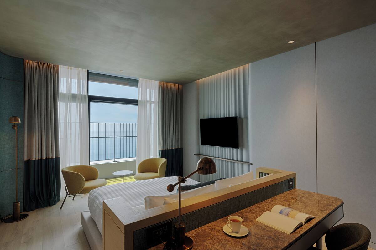 A modern hotel room with a large bed, a desk with a lamp, a chair, a TV mounted on the wall, and a view of the city.
