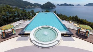Une piscine rectangulaire à débordement, avec un jacuzzi au centre, entourée de chaises longues et de parasols, offrant une vue sur une étendue d’eau et des îles lointaines.