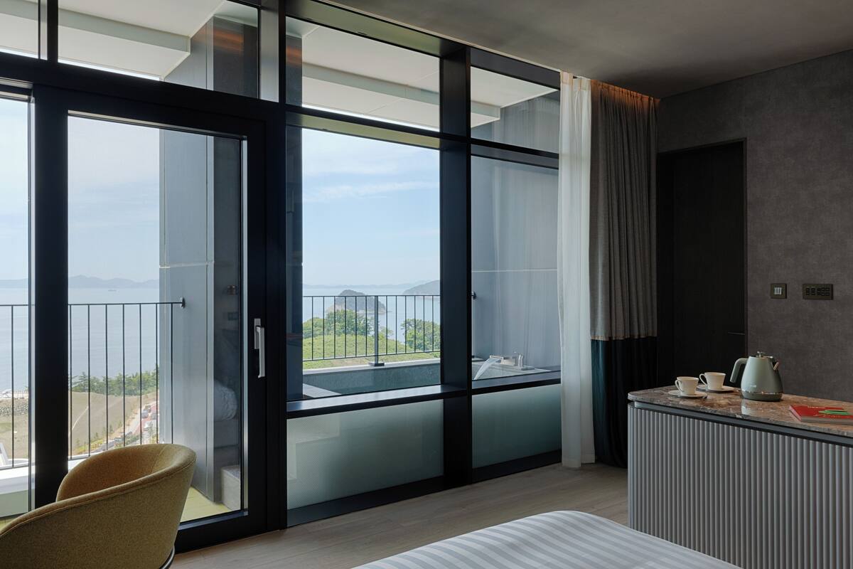 A modern hotel room with a large sliding glass door leading to a balcony with a view of the sea and mountains.
