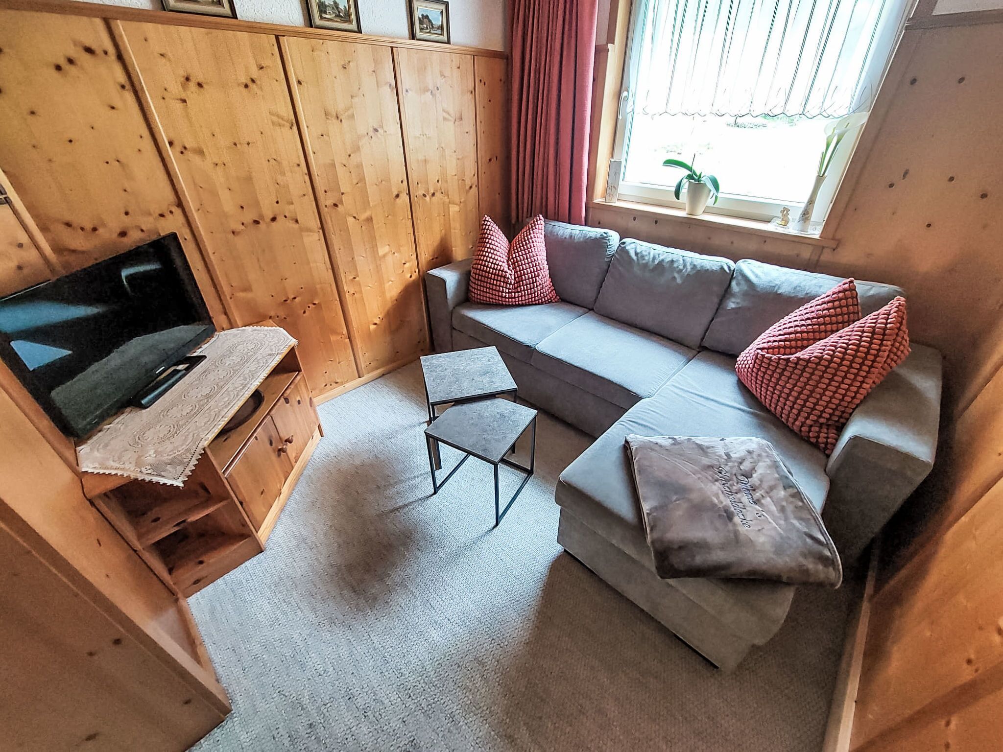 A cozy room with a wooden wardrobe, a grey sofa with red and white checkered cushions, a small table, and a TV on a wooden stand.