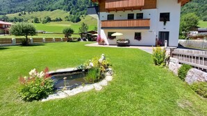 A two-story chalet with a balcony, a small pond, and a mountain backdrop.