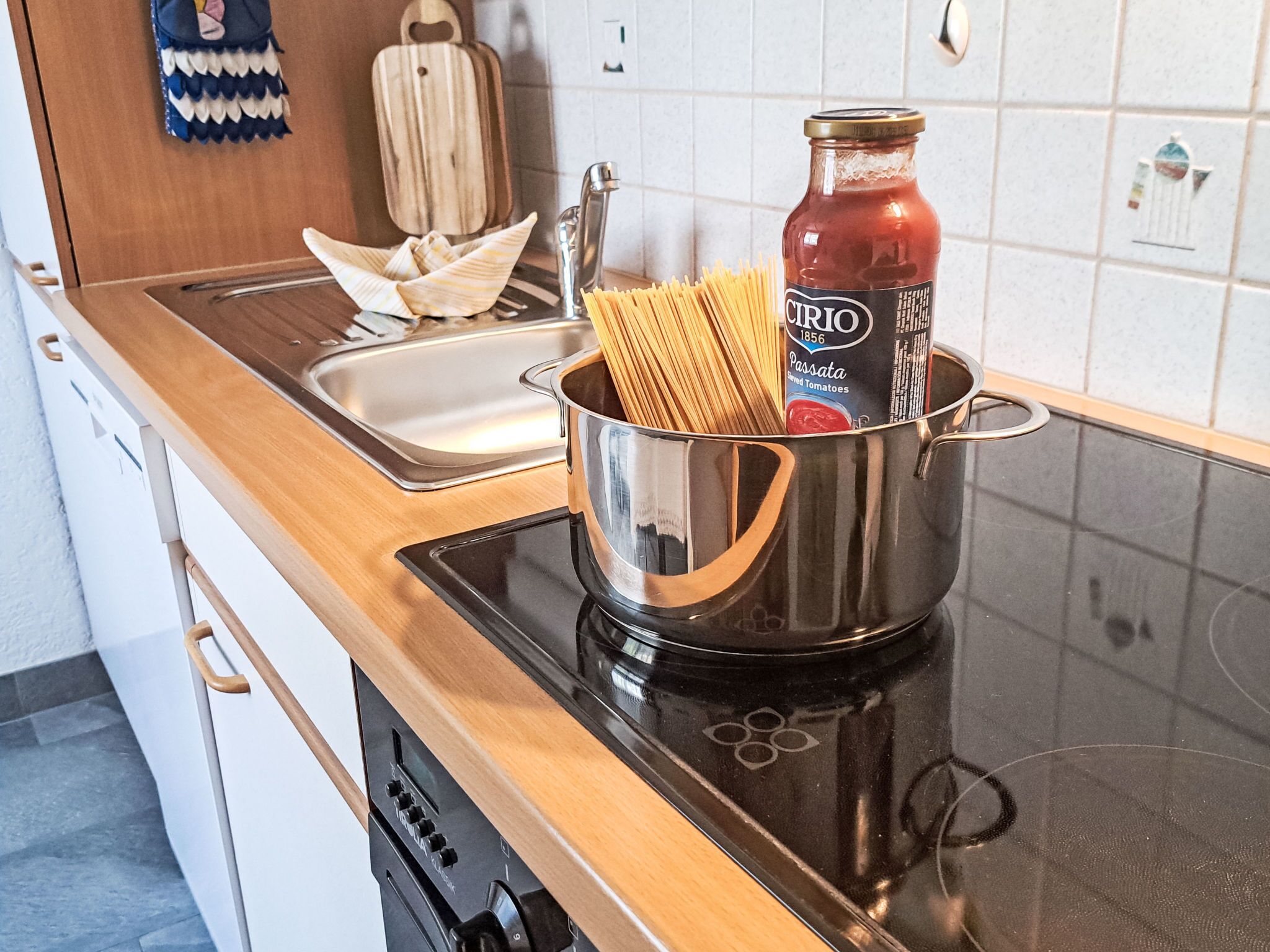 Une cuisine avec une casserole en acier inoxydable sur une plaque de cuisson à induction, contenant des spaghettis et une bouteille de sauce tomate.