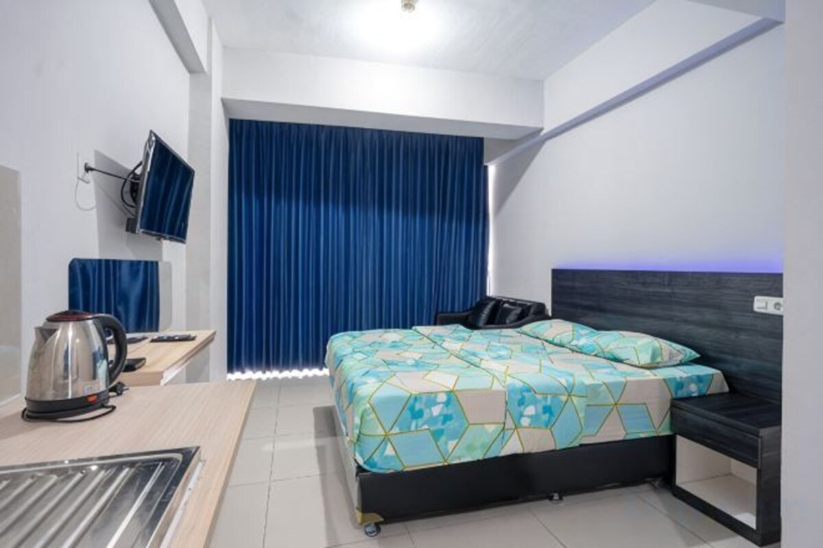 A hotel room with a bed, a desk with a kettle, a chair, a TV, and a curtain.