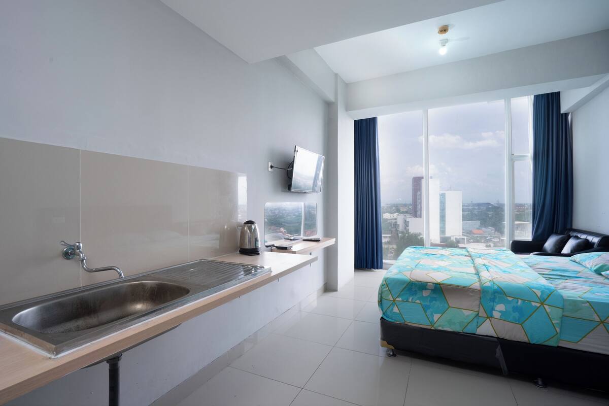 A modern hotel room with a large window, a bed with a geometric-patterned bedspread, a built-in sink, and a flat-screen TV mounted on the wall.