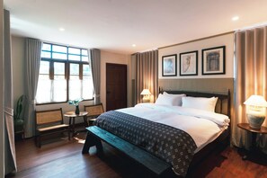 A hotel room with a large bed, a bench, a small table, and a window with curtains.