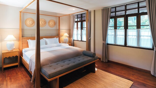 A four-poster bed with a grey upholstered bench, wooden headboard, and a woven rug on a wooden floor.