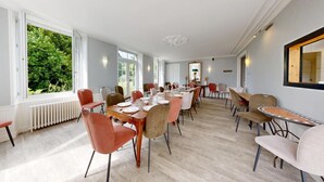 A dining area with multiple tables and chairs, large windows with a view of greenery, and a white ceiling with a central light fixture.
