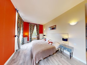 Una habitació d'hotel amb un llit gran, una taula de nit amb una làmpada, un radiador i una finestra amb cortines.