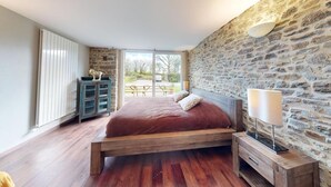 Un dormitorio con pared de piedra, cama de madera, mesita de noche y vistas al exterior.