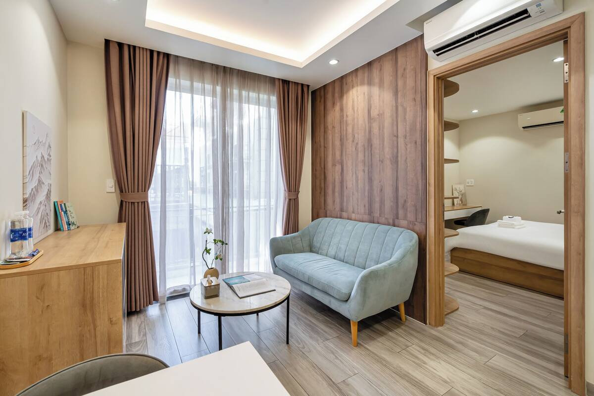 A modern hotel room with a light blue sofa, a wooden desk, a small round table, and a bed with a wooden headboard.
