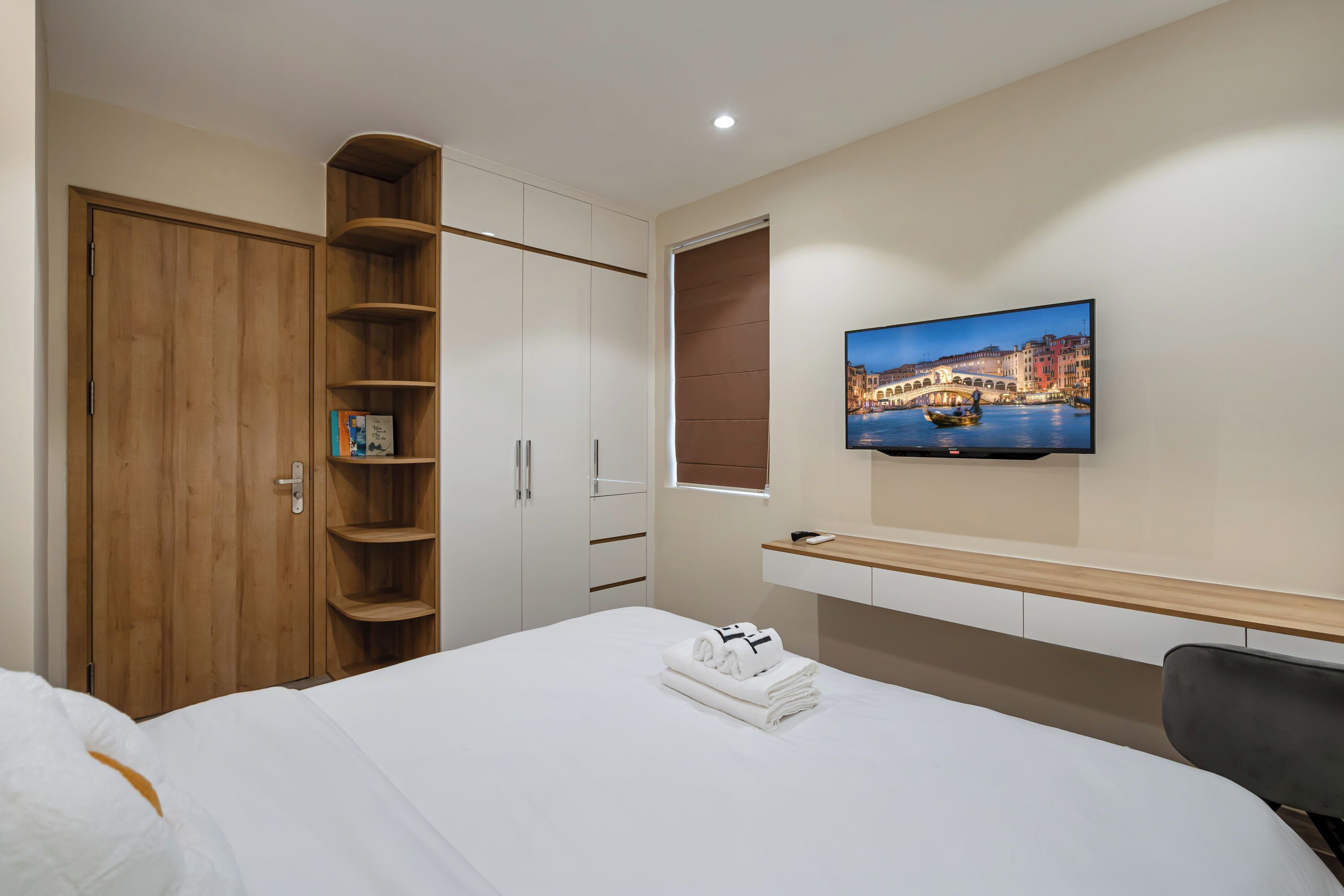 A hotel room with a bed, a wooden wardrobe, a TV mounted on the wall, and a window with a cityscape view.