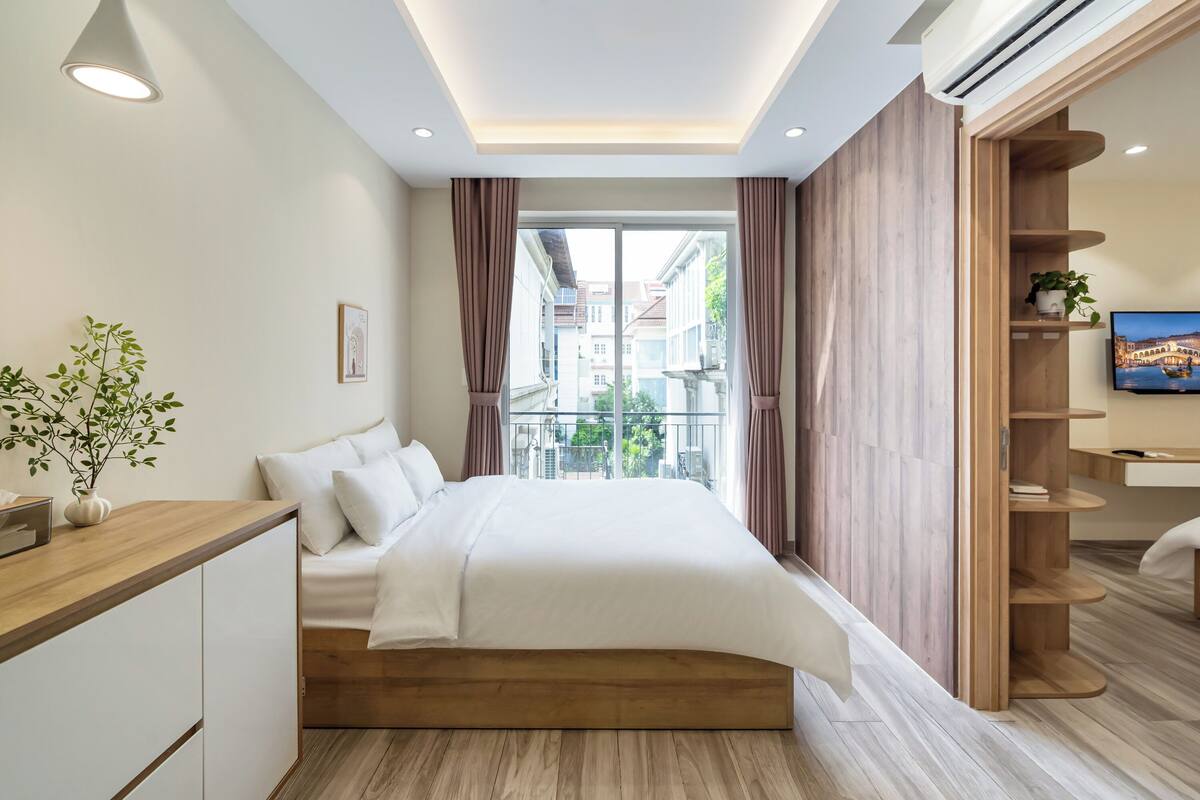 A modern hotel room with a large bed, wooden headboard, a bedside table, a potted plant, a TV, and a balcony with a city view.