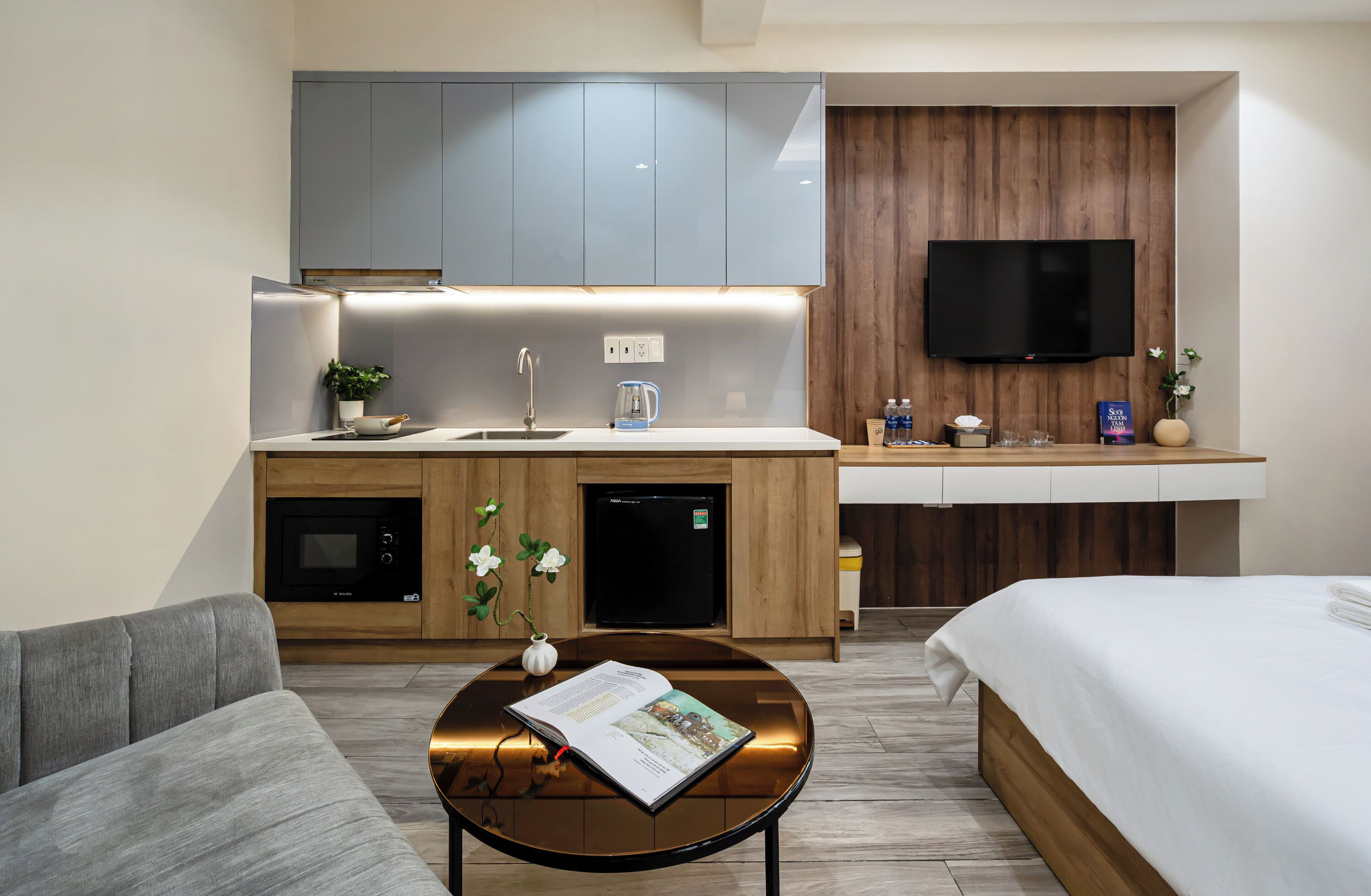 A modern hotel room with a kitchenette, a flat-screen TV, a sofa, and a bed.