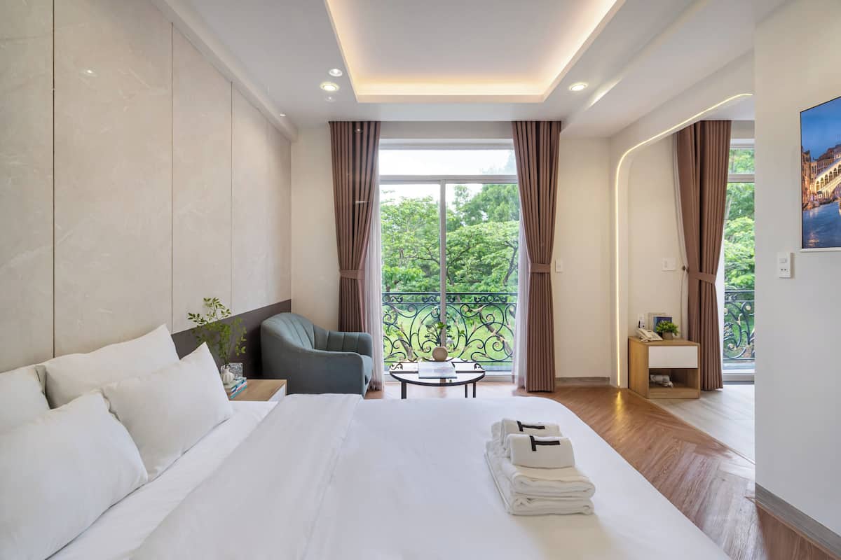 A hotel room with a large bed, a seating area with a table, and a balcony with a view of greenery.