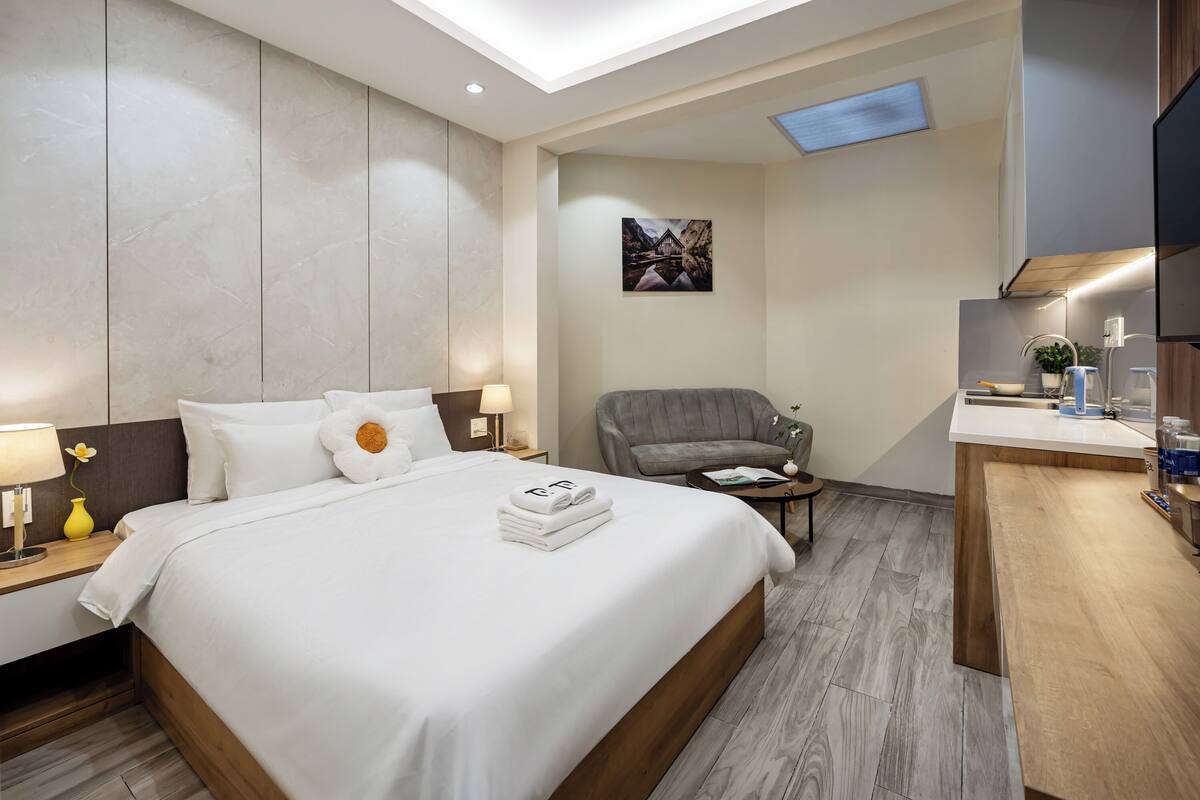A modern hotel room with a large bed, a desk, a chair, and a small kitchenette.