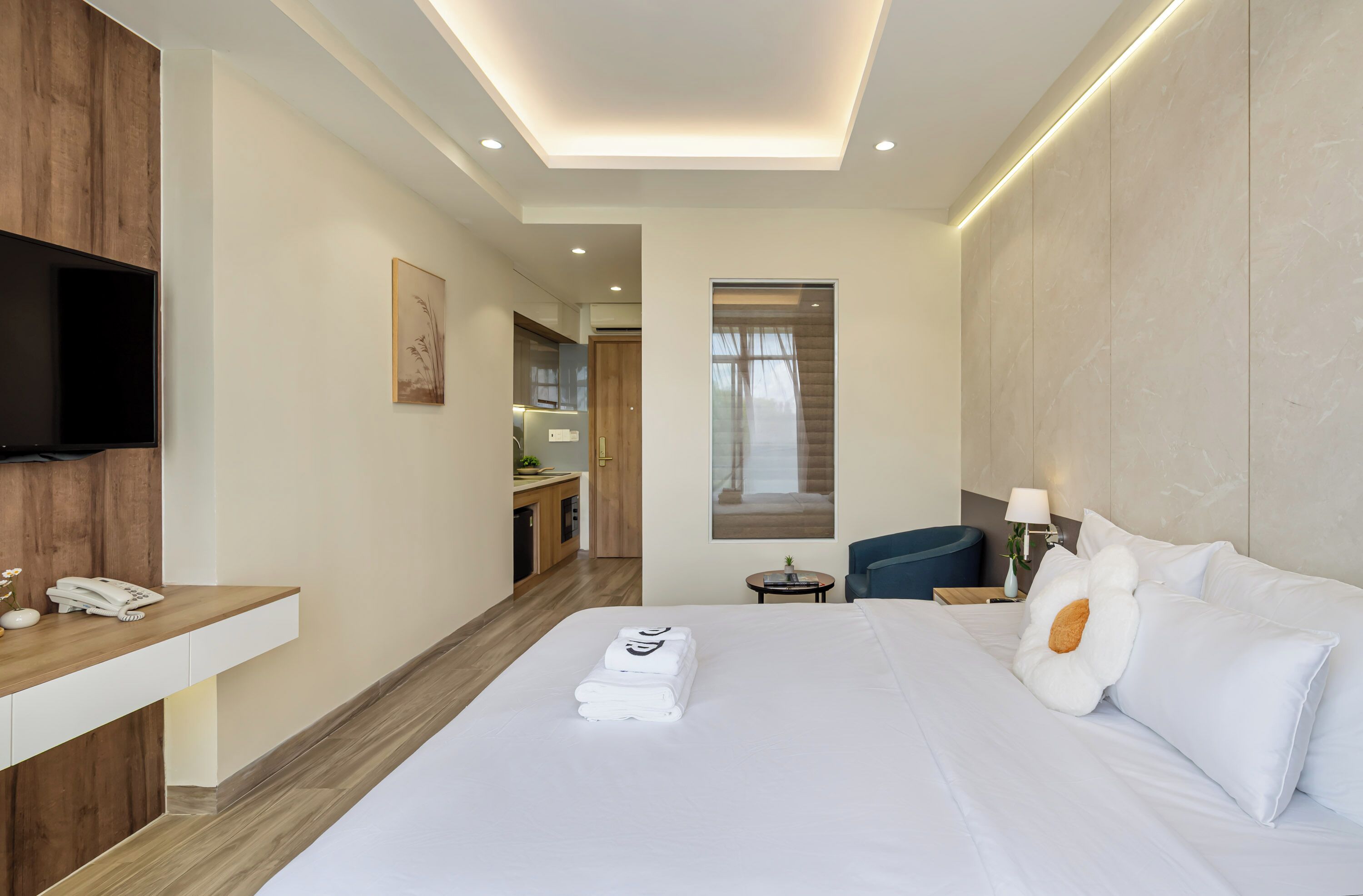 Habitación de hotel con una cama bien hecha, un televisor de pantalla plana montado en la pared, un escritorio pequeño con teléfono y vistas al pasillo y otra habitación.