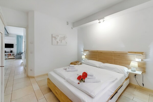 A hotel room with a large bed, a bedside table with a lamp, a world map on the wall, and a view of the adjacent room.