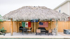 Zona junto a la piscina con techo de paja, banderas y mobiliario exterior.