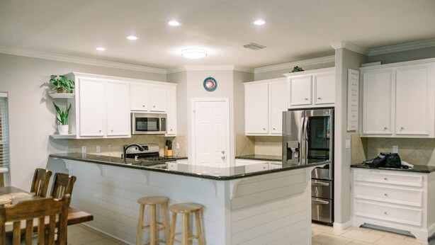 Una cocina moderna con armarios blancos, una isla central con taburetes, electrodomésticos de acero inoxidable y piso de baldosas.