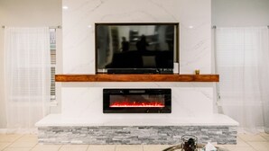 Una sala de estar moderna con un televisor de pantalla plana grande montado en una pared de mármol, una chimenea empotrada debajo y un estante de madera arriba.