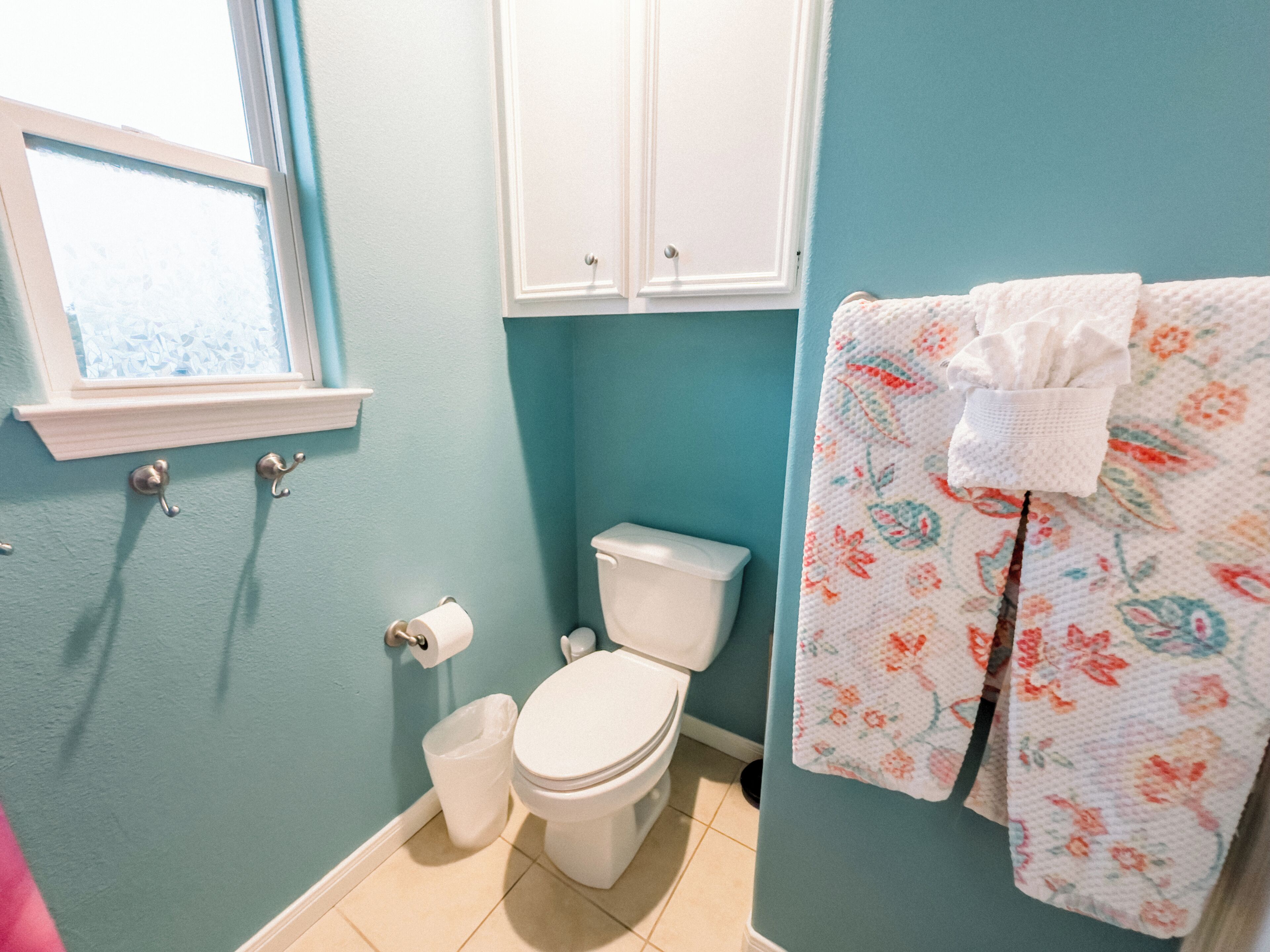 Et badeværelse med toilet, skraldespand, et vindue og en håndklædestang med et blomstret håndklæde.