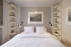 Una camera da letto con un letto, cuscini bianchi, un quadro incorniciato appeso al muro e scaffali a incasso con diversi oggetti.
