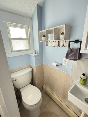 A small bathroom with a toilet, a window, a shelf with toiletries, and a towel rack. - Charming 2-bedroom building in delightful Hull with ocean view (Hull)