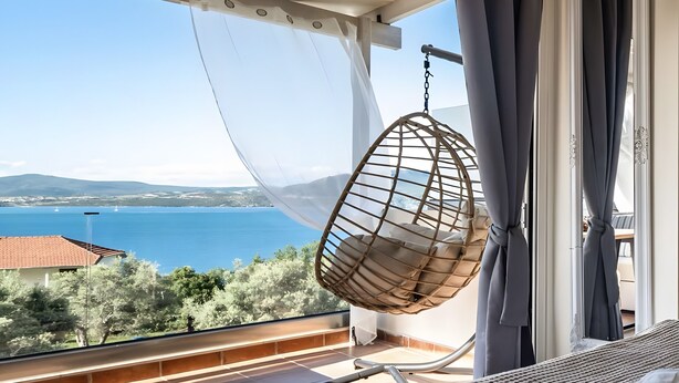 A balcony with a hanging chair, a bed with a white towel, and a view of the sea and hills.