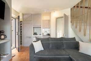 Una sala de estar moderna con un sofá gris, una cafetera y una zona de cocina con armarios blancos y una mesa de comedor.