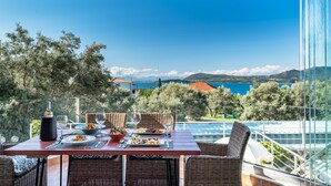 A dining table with plates of food, glasses, and a bottle, set on a balcony with a view of trees and mountains.