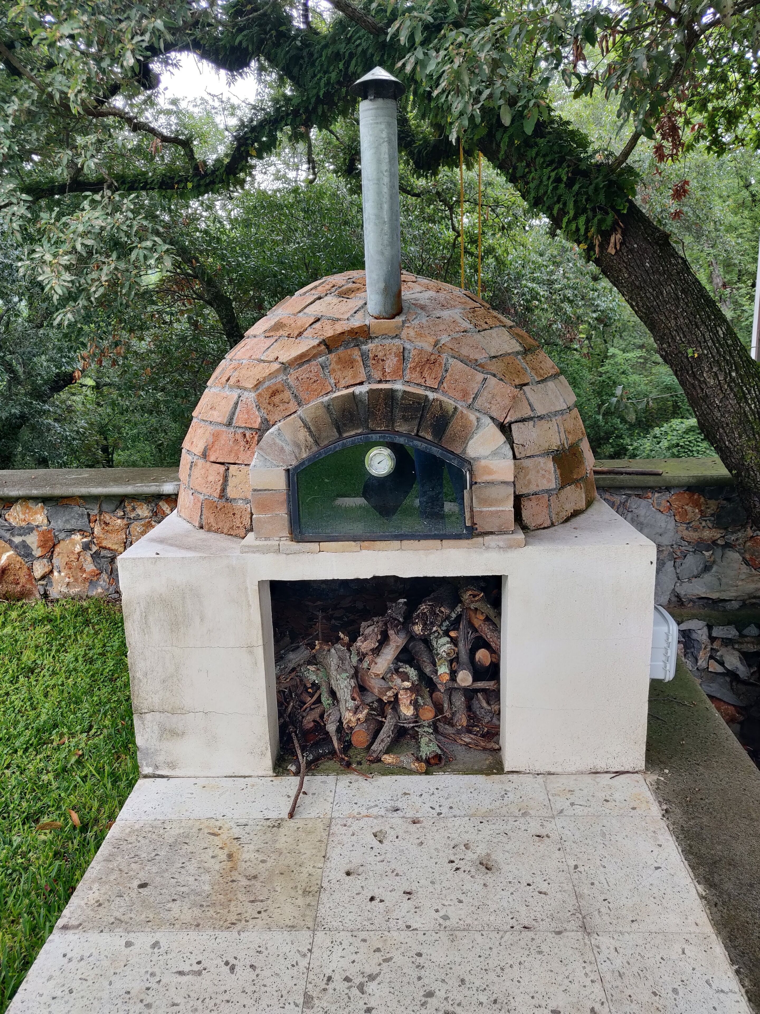 Un four à pizza en briques installé à l’extérieur, doté d’une cheminée, et entouré de verdure et d’un mur en pierre.