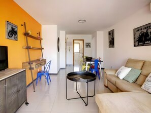 Un salon comprenant un canapé, un téléviseur, un coin repas avec des chaises et un coin cuisine avec un réfrigérateur.