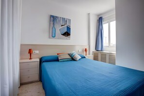 Ein Schlafzimmer mit einem Bett, einem Nachttisch, einer Lampe, einem Fenster mit Vorhängen und einem Wandbild.