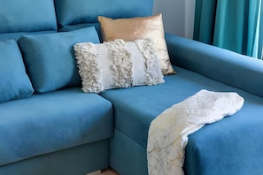 Ein blaues Sofa mit drei Kissen, davon eines golden und zwei weiße mit Fransen.