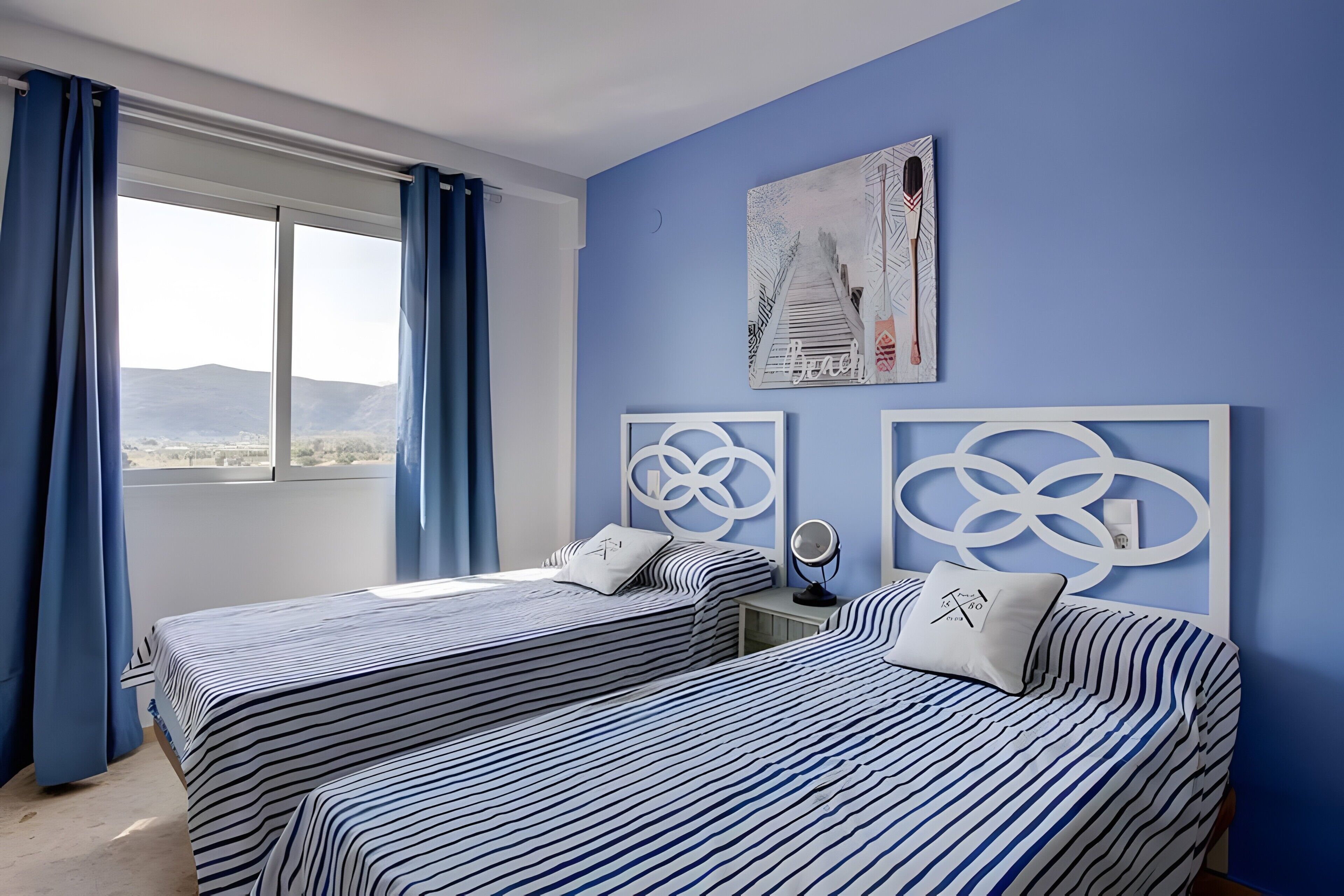A room with two beds, a window with curtains, a wall with artwork, and a nightstand with a mirror.