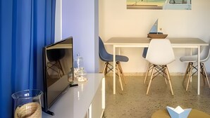 A modern hotel room with a white desk, two chairs, a small table, a TV, and a painting of a boat on a beach.