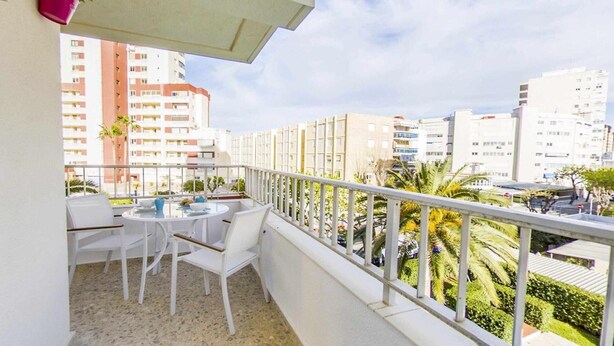A balcony with a table and chairs, a railing, and a view of surrounding buildings and greenery.