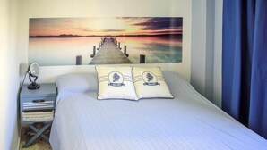 A bedroom with a bed, pillows, a nightstand, a lamp, and a large wall painting of a pier.
