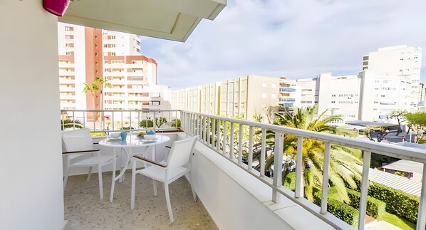 A balcony with a white table and chairs, a potted plant, and a view of surrounding buildings and greenery.