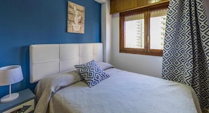 A bedroom with a bed, a bedside table with a lamp, a window with patterned curtains, and a wall art piece.