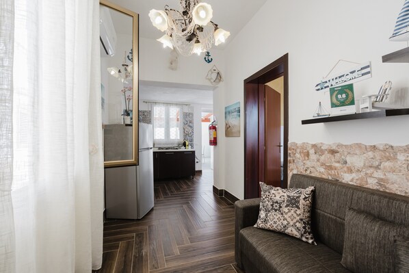 A hotel lobby with a wooden floor, a sofa with a patterned pillow, a chandelier, a kitchen area, and a stone wall.