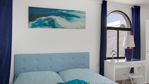 Un dormitorio con cama, escritorio, silla, ventana con cortinas y un cuadro del océano.