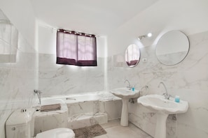 A bathroom with a bathtub, two sinks, a toilet, and a window with curtains.