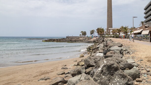 Een kustgezicht met een rotsachtig strand, een hoge toren en een verharde promenade met palmbomen.